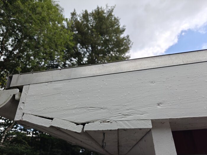 Närbild av vitmålad takfot med metallbeklädnad ovanpå; tecken på slitage och fukt, omgivet av träd och himmel i bakgrunden.