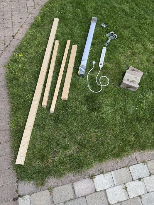 Virke, lysrör i förpackning, grenuttag med sladd, hjul och murstenar ligger på en gräsmatta, redo för DIY-renoveringsprojekt.