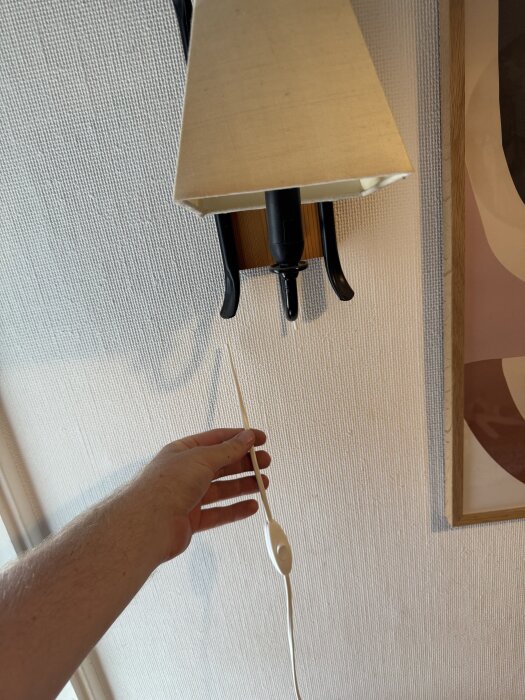 En hand håller i en vit lampsladd som är ansluten till en vägglampa med beige skärm, monterad på en ljus mönstrad vägg.