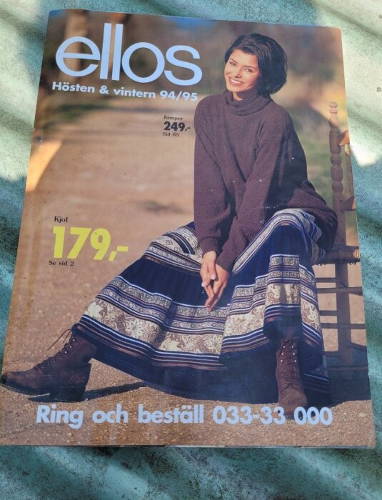Ellos-katalog från höst och vinter 94/95 med kvinna i brun tröja och mönstrad kjol, sittande och leende. Telefonnummer för beställning syns.