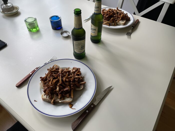 Två tallrikar med bröd toppat med stekta kantareller på ett bord, omgivna av bestick, flaskor och färgade glasljus.