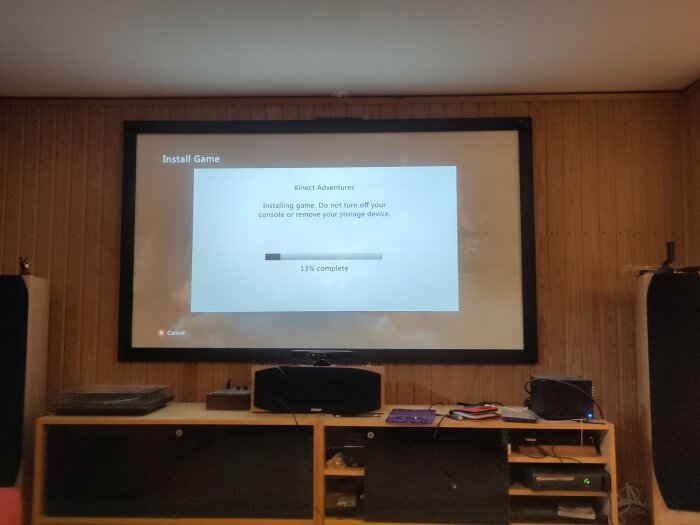 Installationsskärm för spelet Kinect Adventures på en TV, med framstegsindikator som visar 13% klart, i ett rum med trämönstrad vägg och högtalare.