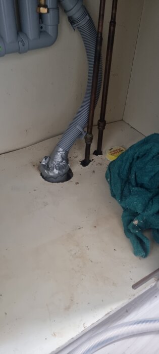 Rör under diskbänk med temporär silvertejpstätning, fuktskadad yta, och grönt tygstycke.