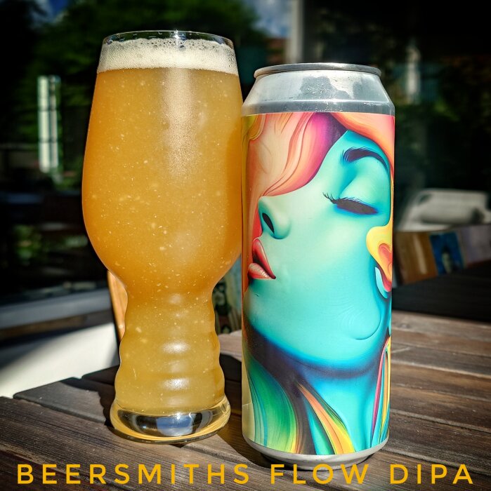 Ett glas fyllt med Beersmiths Flow DIPA bredvid en färgglad burk med konstverk på ett bord utomhus i solen.
