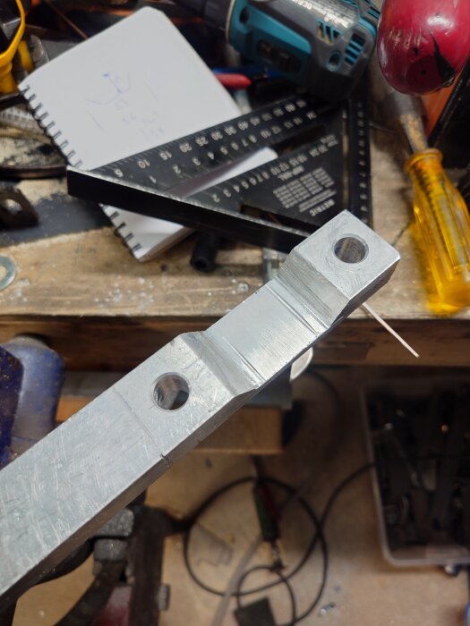 En metallbit med förborrade hål på en arbetsbänk, omgiven av verktyg som en skruvmejsel och linjal, samt en skiss på ett block.