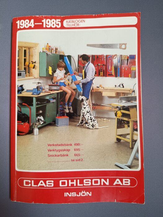 Omslag av Clas Ohlson-katalog 1984-1985 med verkstadsmiljö, pojke och man vid arbetsbänk, dalmatiner på golvet, verktyg på väggen.