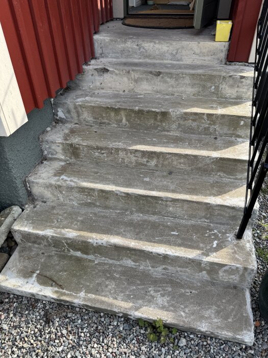 Betongtrappa vid entré med avflagad yta där tidigare klinker tagits bort, omgiven av röda plåtväggar och grus på marken nedanför.