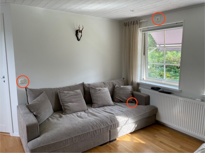 Grå soffa vid vit vägg med dödskalle på. Ovanför soffan finns strömställare och vägguttag, fönster till höger. Rödmarkerade elkomponenter.