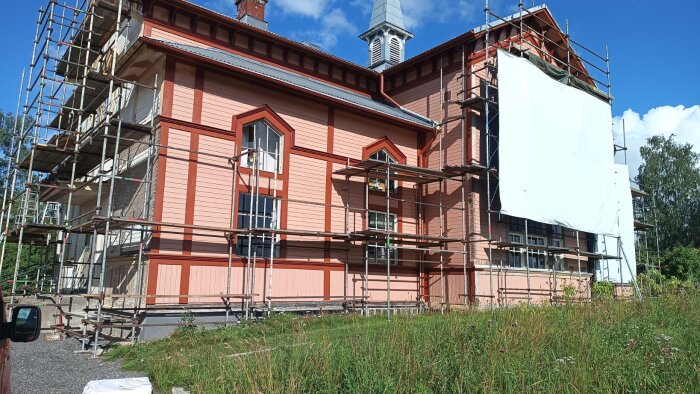 Stort hus med pågående renovering av fasaden, byggställningar runt om, delvis täckt med vit plast, blå himmel i bakgrunden.