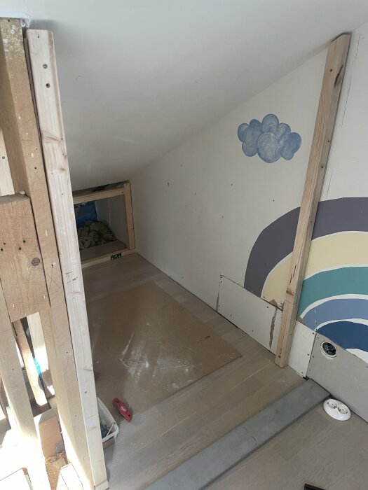 Under en ombyggnad med träreglar och gipsskivor, syns en vägg med en målad regnbåge och moln. Verktyg och material ligger på golvet.