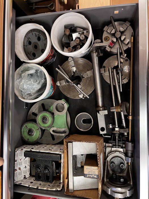 Verktyg och maskindelar organiserade i låda, inklusive kugghjul, chuckar och borrtillbehör.