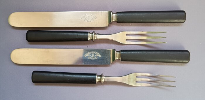 Bestick i kolstål från Eskilstuna jernmanufaktur med svärtade päronträhandtag, inklusive knivar och gafflar, på en lila yta.