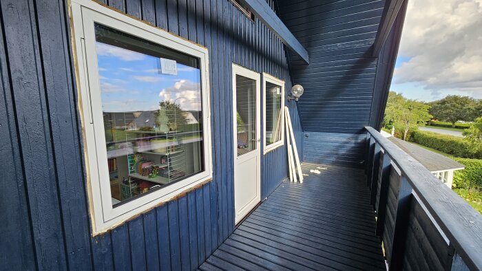 Nya fönster på andra våningen i en blåmålad träpanel, med synliga glipor runtom, på en balkong med räcke och trädgård i bakgrunden.
