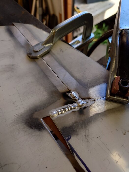 Svetsad aluminiumplåt fastsatt med klämma, visar ojämna svetsfogade skarvar. Verkar vara del av instrumentpanelprojekt, enligt text från inlägget.