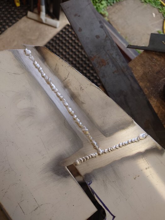 Svetsad aluminiumplåt med grova svetsar längs en kant, på ett arbetsbord i en verkstadsmiljö.