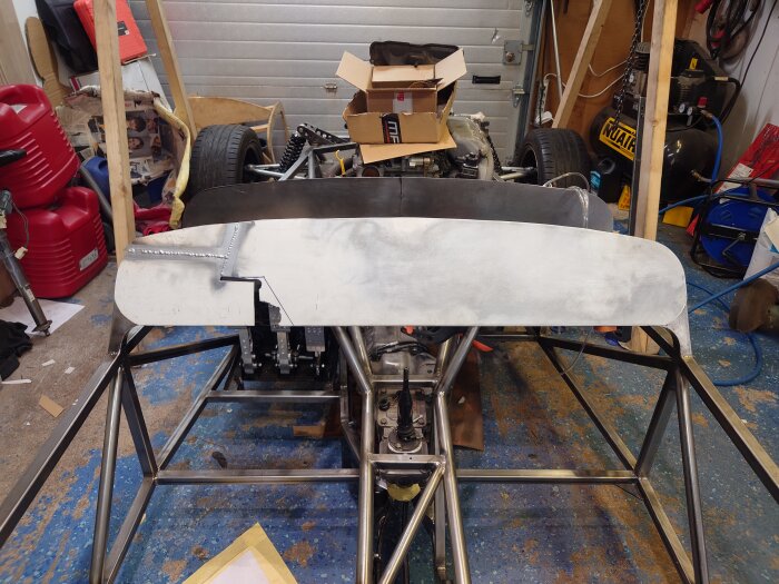 Bilram med hemmabyggd instrumentpanel i aluminium och synliga svetsfogar i en verkstad med verktyg och kartonger i bakgrunden.