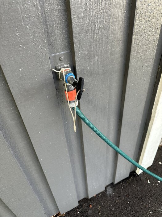 Frostsäkert vattenutkast med avstängd ventil och grön trädgårdsslang fäst vid en grå trävägg.