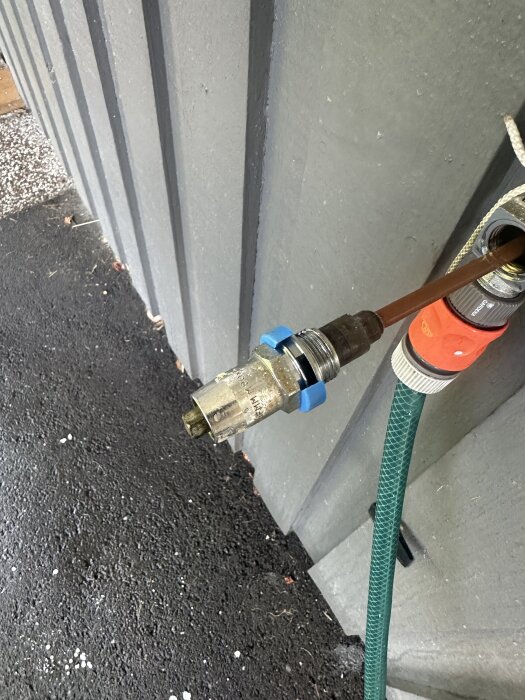 Frostsäkert vattenutkast monterat på en husvägg, med avskruvat rör och fästmuttrar synliga. Grön trädgårdsslang kopplad till utkastet.