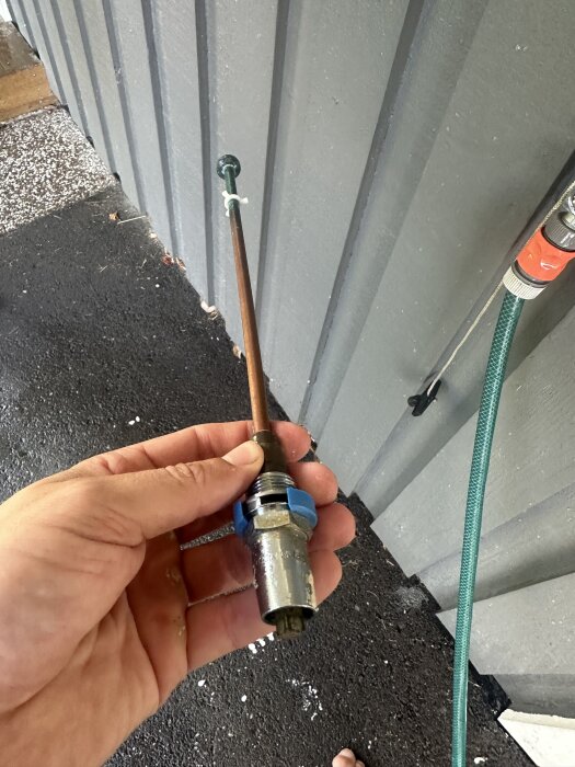 Hand som håller ett frostsäkert vattenutkast mot en grå vägg, synlig trädgårdsslang kopplad bredvid.