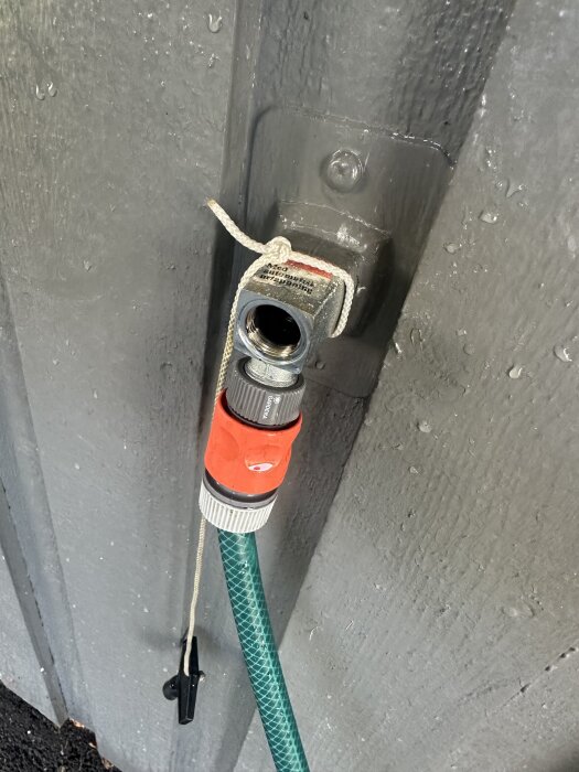 Frostsäkert vattenutkast med borttaget munstycke och ansluten trädgårdsslang, knutet med ett snöre på en våt vägg.