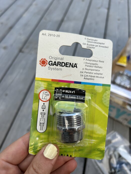 Förpackning med Gardena-adapter för vattenslang, Art. 2910-20, hålls i hand med ljusgul nagellack, uteplats av trä i bakgrunden.