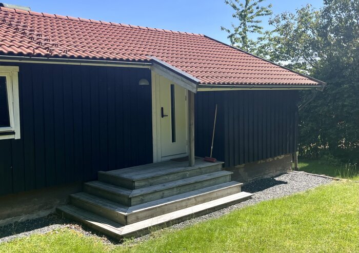 Nybyggd entrétrappa i trä med fyra steg utanför ett hus med rött tegeltak, inramad av gräsmatta och grus.