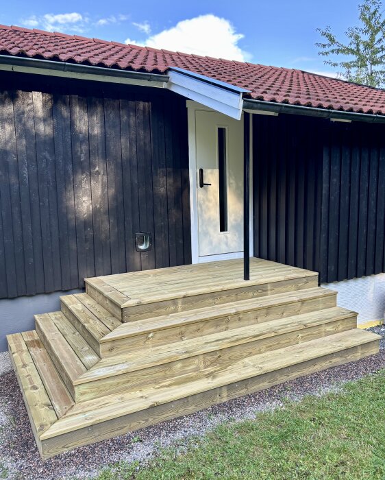 Nybyggd entrétrappa i trä med stålstolpe, byggd med 174x45 reglar och 145x28 trall. Husets fasad i mörkt trä och tegelrött tak.