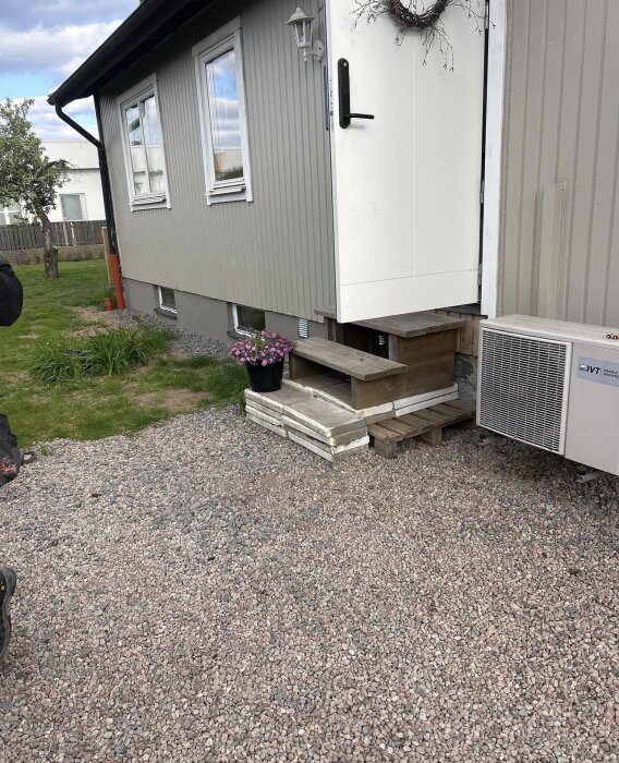 Entré med trappsteg i trä mot en putsad husvägg, blomkruka bredvid och grus på marken.