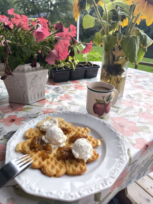 Våfflor med hjortronsylt och grädde på en vit tallrik, omgiven av blommor och en kopp kaffe på ett blommigt bord.