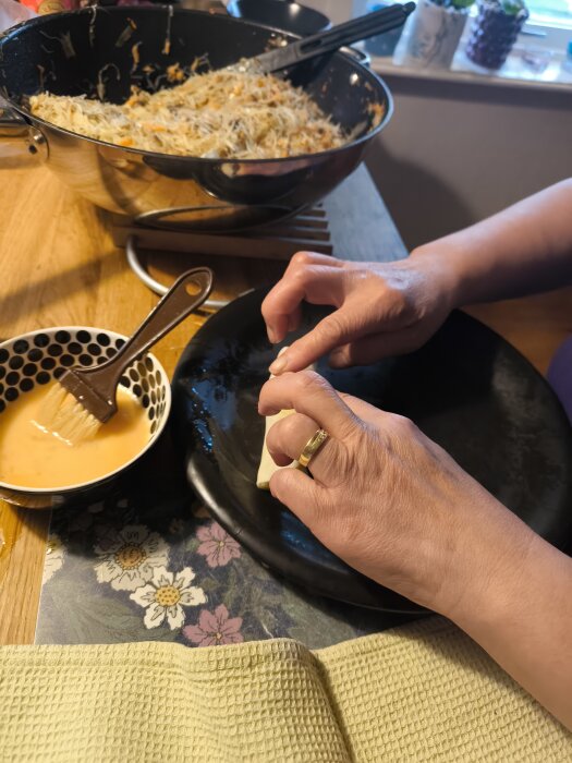 Person rullar vårrullar vid köksbord, omgiven av ingredienser och en skål med äggblandning.