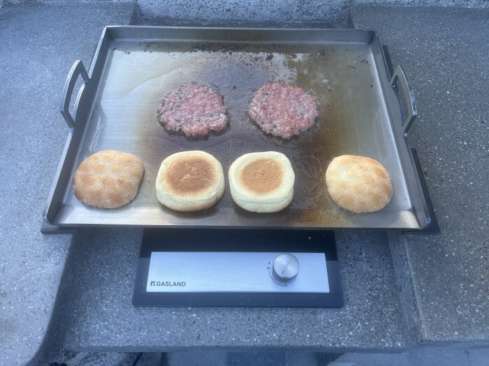 Stekhäll med två smashburgare och hamburgerbröd under tillagning i ett utekök.