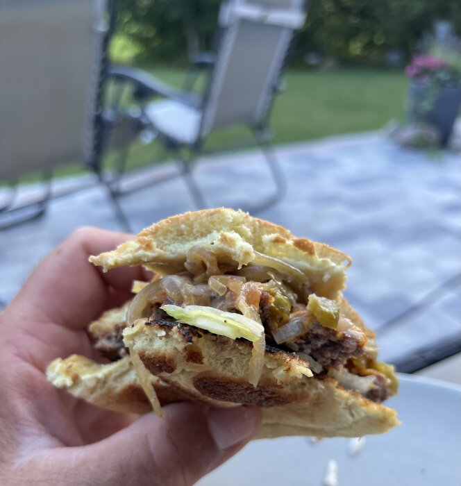Smashburgare med sallad och lök i närbild, hålls i handen. Bakgrunden visar utemiljö med stolar och gräsmatta.