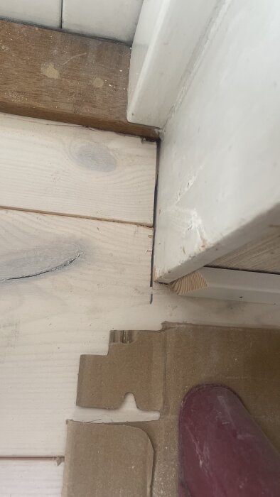 Massivt furugolv med ojämna glipor och skador nära en vägglist och rör i ett vardagsrum under renovering.