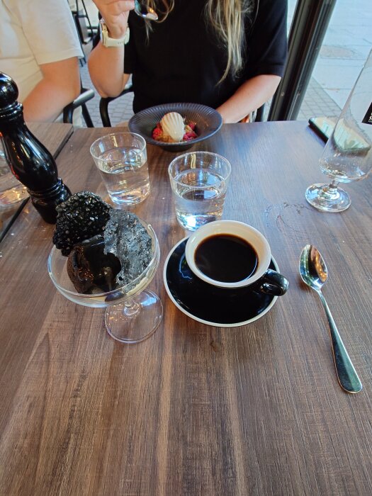 Svart kaffe i kopp, lakritsparfait med citronglass i skål, två glas vatten, vinflaska, pepparkvarn, dessert i skål med jordgubb och grädde på träbord.