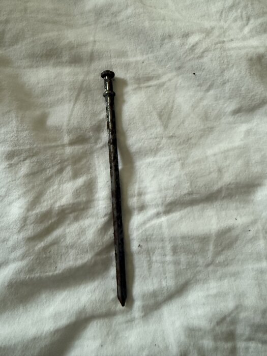 Gammalt järnspett eller stång med rund huvudet och spetsig ände, liggande på ett vitt tyg.