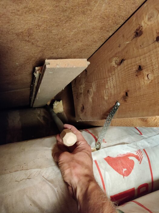 Hand håller i en ihopsatt armeringsjärnsskruv med knopp, ovanför isolering och träbjälkar i en vind.