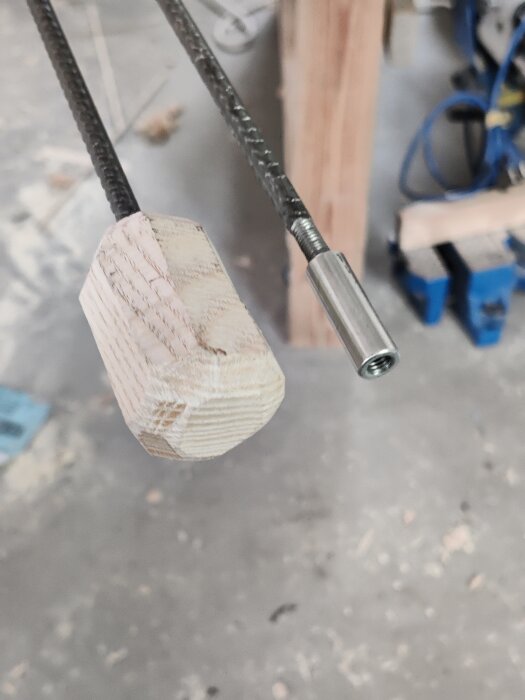 Närbild på två armeringsjärn skruvade ihop med en metallskarvmuff och en träknopp fäst i änden av det ena, på cementgolv i verkstad.