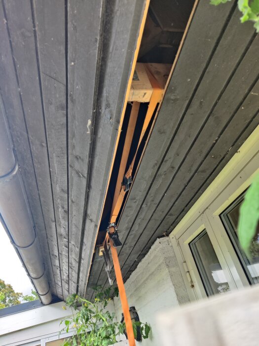 Utsikt under takfot där panel saknas, avslöjande trästruktur. Orange spännband hänger för att underlätta tillgång vid installation av kamera.
