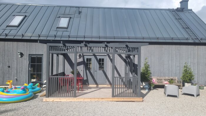 En nybyggd gråmålad terrass med stolpar och räcke framför ett hus, omgiven av utemöbler och lekutrustning.