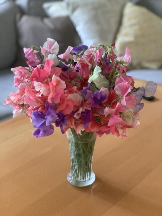 En vas med färgglada luktärter i rosa, lila och vita färger står på ett träbord med en soffa i bakgrunden.
