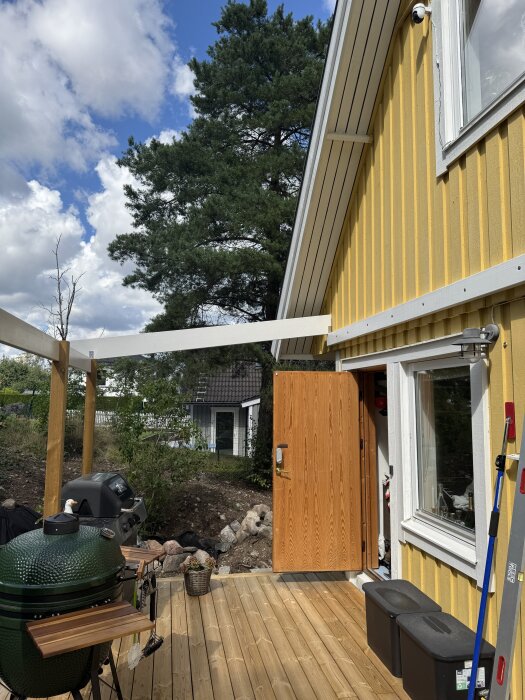 Altan med trädäck under konstruktion, trästolpar står redo för skärmtak av kanalplast, och en grill ses nedanför ett gult hus med lockpanel.