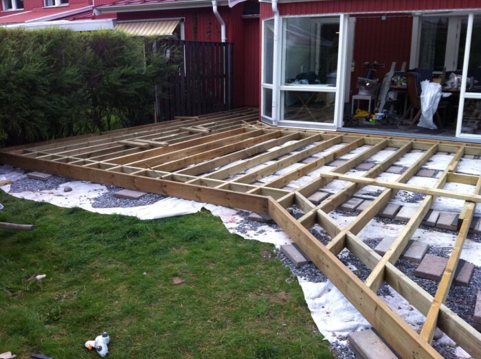 Trädäck under konstruktion, träreglar uppställda på plattor vid husvägg, förberett för växthus eller altan, omgiven av gräsmatta och buskar.