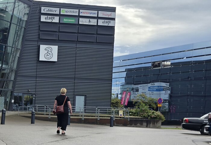Kvinna går mot en modern byggnad med olika butiksskyltar, inklusive Glitter, Apoteket och Intersport. Reflekterande glasfasad syns i bakgrunden.