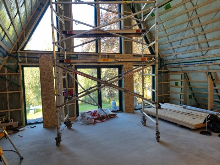 Ett rum under renovering med stora fönster, byggställning, isolerade väggar och reglar, samt byggmaterial på betonggolvet.