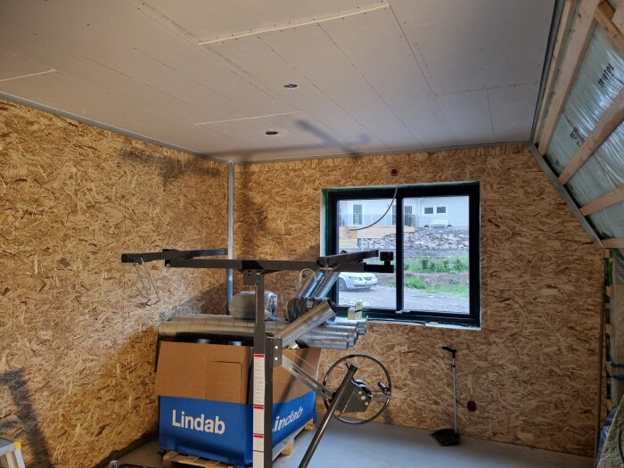 Sovrum med gipstak och träväggar. Fönster med utsikt över trädgård. Kartonger och byggmaterial på golvet.