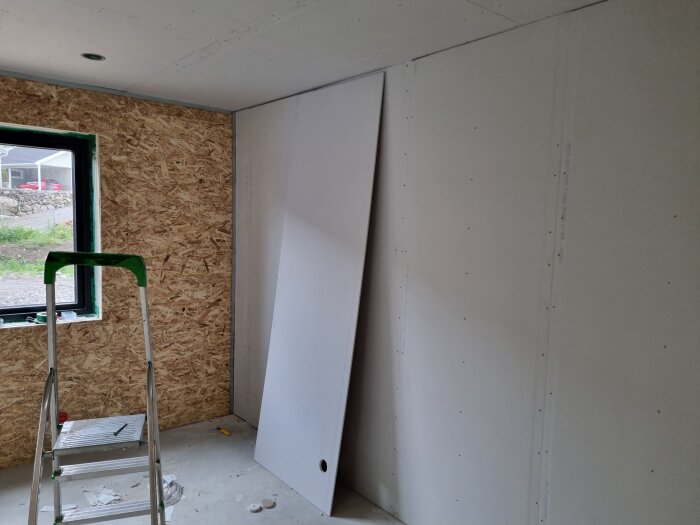 Sovrum med osb-vägg, gipstak och gipsad vägg, stege nära fönster, och en stor gipsskiva lutad mot väggen.