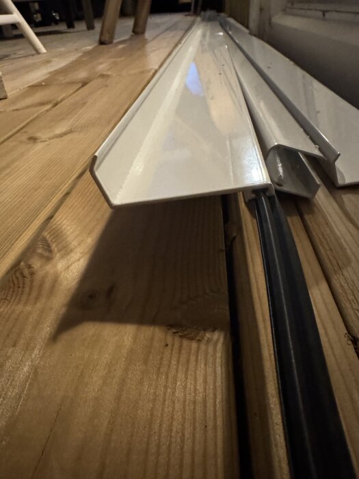 Vit vägganslutningsprofil av metall liggande på en träyta, avsedd för montering av skärmtak.