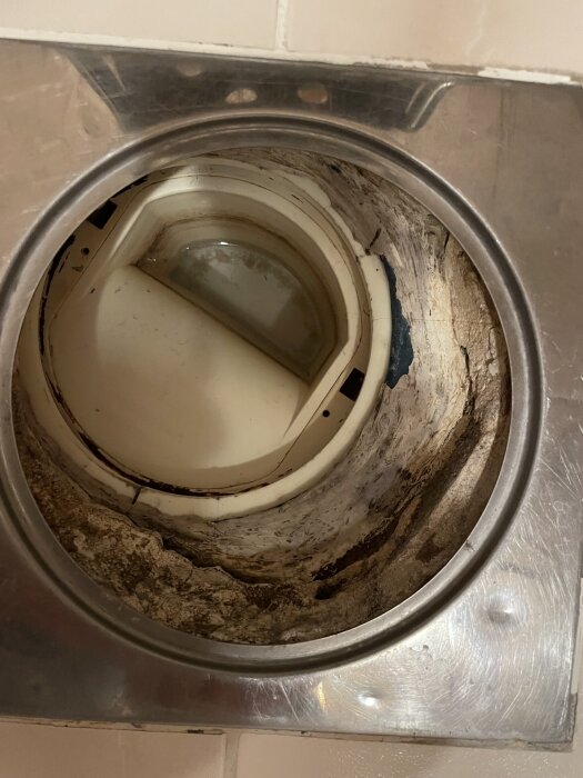 Bild på en golvbrunn i ett duschutrymme, med vit plastvattenlås, klämring och blå våtrumsmatta, omgiven av kakel och gjutet material.