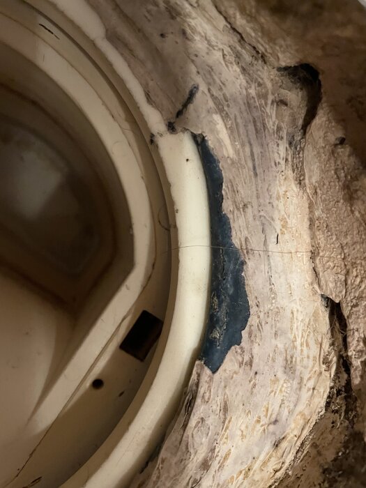 Närbild av en golvbrunn i ett duschutrymme, visar vit plast vattenlås, blå våtrumsmatta synlig under gjutet material, tecken på ålder och slitage.