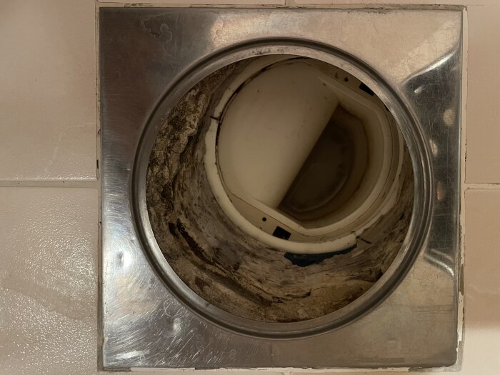 Golvbrunn i dusch med vit plast vattenlås, blå våtrumsmatta och kromring, omgärdad av kakel. Risk för vattenläckage påpekat.